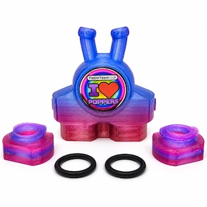 Könnte beinhalten: Ein 3D-gedrucktes Gerät mit blau-rosa Farbverlauf, zwei oberen Vorsprüngen und einem zentralen runden Aufkleber mit der Aufschrift "PopperTopper.co.uk I LOVE POPPERS" und einem roten Herz. Zwei passende Basisteile und zwei schwarze O-Ringe sind enthalten.