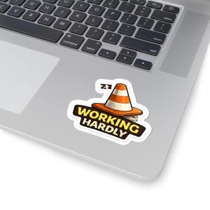 May include: A die-cut sticker featuring an orange and white traffic cone resting on a dark brown base with the text WORKING HARDLY in bold yellow capital letters. The sticker is placed on the silver surface of a laptop keyboard.