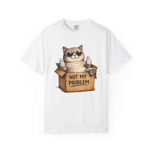 Puede incluir: Una camiseta blanca con el gráfico de un gato marrón claro con gafas de sol negras, sentado dentro de una caja de cartón marrón. El gato muestra ambos pulgares hacia arriba y el texto en el frente de la caja dice NOT MY PROBLEM en negro.