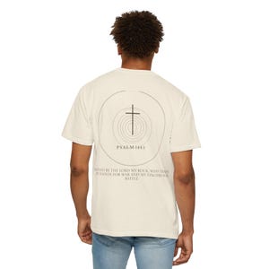 Op de afbeelding: Een off-white t-shirt met korte mouwen en een zwarte afbeelding op de achterkant. Het ontwerp bevat een centraal kruis binnen concentrische cirkels, de tekst PSALM 144:1 en het vers: BLESSED BE THE LORD MY ROCK, WHO TRAINS MY HANDS FOR WAR AND MY FINGERS FOR BATTLE.