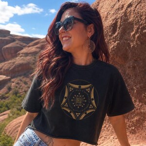 May include: A black cropped t-shirt featuring a circular gold graphic design on the chest. The graphic consists of a central starburst pattern surrounded by geometric shapes, thin lines, and small dots, all enclosed within a circular border.
