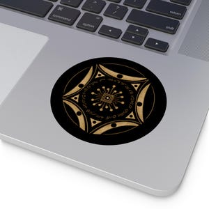 Puede incluir: Una pegatina circular con fondo negro y un diseño geométrico de color dorado. El centro muestra un cuadrado con líneas radiantes, rodeado por un anillo de símbolos y un borde exterior de formas puntiagudas con pequeños círculos negros.