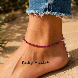 May include: An anklet featuring small, round, deep pink ruby beads strung on a delicate chain. The piece includes a silver-toned metal extender chain finished with a small heart-shaped charm. The text "Ruby Anklet" is visible on the image.