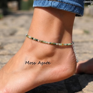 May include: An anklet featuring small, round, multi-tonal green moss agate beads strung together. The piece includes a silver-toned metal chain extender with a small bead at the end. The text "Moss Agate" is visible near the ankle, with branding above.