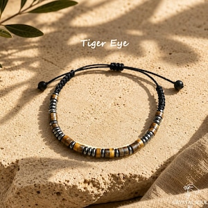May include: A bracelet featuring alternating cylindrical tiger eye beads and small metallic gray spacer beads on a black cord. The cord has an adjustable sliding knot closure with two small black beads at the ends. The text Tiger Eye is visible.