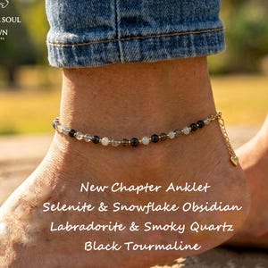 May include: An anklet featuring a delicate gold-tone chain strung with small, alternating beads of selenite, snowflake obsidian, labradorite, smoky quartz, and black tourmaline. A small gold-tone heart charm hangs from the end of the chain.