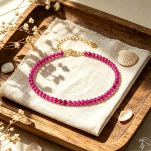 May include: A delicate bracelet featuring small, faceted, vibrant magenta-pink gemstone beads arranged in a circular pattern. The jewelry piece includes a thin gold-toned metal chain with a lobster clasp closure and an adjustable extender link.