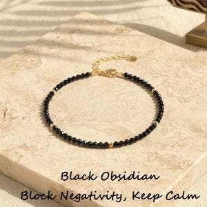 Puede incluir: Una pulsera delicada con pequeñas cuentas de obsidiana negra facetadas en una cadena dorada con cierre de mosquetón. El texto "Black Obsidian Block Negativity, Keep Calm" aparece debajo de la joya y un letrero de madera dice "CrystalSoulByShawn".
