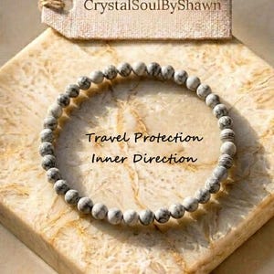 May include: A circular bracelet made of small, round, white and grey marbled beads. A rectangular beige tag is positioned above the bracelet with the text CrystalSoulByShawn. The text Travel Protection Inner Direction is centered inside the bracelet.