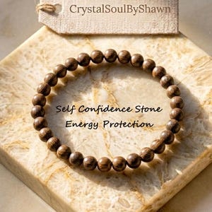 May include: A circular bracelet made of small, round, dark brown wooden beads. A beige rectangular tag above the bracelet displays the text CrystalSoulByShawn. The text Self Confidence Stone Energy Protection is printed in the center of the bracelet.