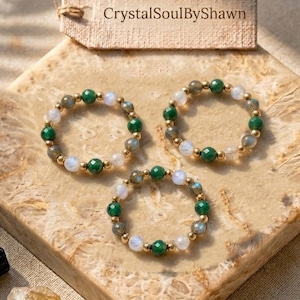 May include: Three beaded rings featuring alternating green and translucent white faceted beads on a thin gold-toned wire. A rectangular tan tag with the text CrystalSoulByShawn is positioned above the rings. A small black stone and a yellow crystal are nearby.