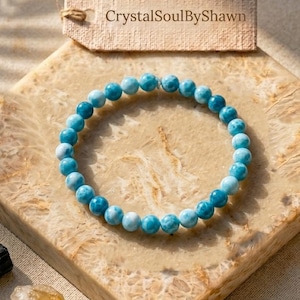 May include: A circular bracelet composed of small, polished, round blue and white Larimar beads. Above the bracelet sits a rectangular beige paper tag with the text CrystalSoulByShawn printed in dark brown letters on the surface.