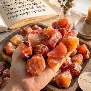 May include: A collection of raw, translucent orange and red carnelian stones held in a palm, with more stones on a wooden tray. An open book displays text about carnelian benefits, and a yellow beeswax candle sits in a ceramic holder nearby.