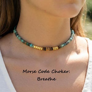 May include: A choker necklace featuring small, round turquoise and brown beads interspersed with metallic gold-tone cylindrical spacers. The text Morse Code Choker: Breathe is printed below the necklace. The beads form a pattern along the neck.
