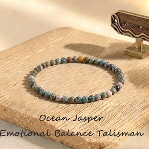 May include: A circular bracelet composed of small, round, multi-colored ocean jasper beads in shades of green, grey, and tan. The text "Ocean Jasper Emotional Balance Talisman" is printed below the bracelet, and a wooden sign reads "CrystalSoulByShawn".