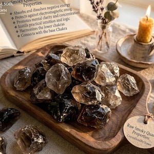 May include: A collection of raw smoky quartz crystals piled on a wooden tray. A circular tag reads Raw Smoky Quartz Properties: CrystalSoulByShawn. An open book displays text about metaphysical properties, and a yellow candle sits nearby.