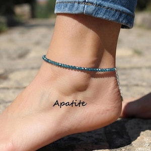 May include: A delicate anklet featuring small, faceted, deep blue apatite gemstone beads. The jewelry piece includes a silver-toned metal chain extension with a clasp, and the word Apatite is printed in black text near the ankle.