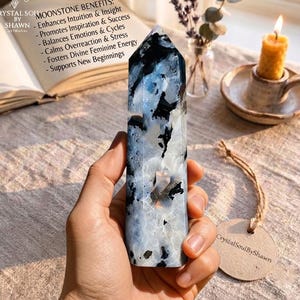 May include: A polished moonstone tower with a hexagonal point, featuring a marbled pattern of translucent white, grey, and deep black inclusions. The stone has a smooth, reflective surface and is held upright, displaying its natural crystalline structure.