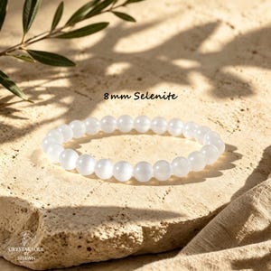 May include: A stretch bracelet composed of smooth, round, translucent white 8 mm Selenite beads. The text 8 mm Selenite is printed above the bracelet, and the logo Crystal Soul by Shawn is printed in the bottom left corner of the image.