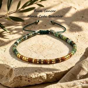 May include: A Morse code bracelet featuring green and brown disc beads with gold-tone spacers, finished with a dark green adjustable cord. The text "Fuck Cancer Morse Code Bracelet" is visible above the jewelry piece in a black script font.