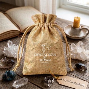 Pu&ograve; includere: Un sacchetto in juta dorata con coulisse, caratterizzato da una grafica bianca di una mano che regge un cristallo e dal testo "CRYSTAL SOUL BY SHAWN ARTISANAL" stampato sul davanti. Il sacchetto ha cordini dorati abbinati con pietre blu scuro.