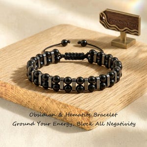 Könnte beinhalten: Ein zweireihiges Armband mit abwechselnden schwarzen runden Perlen und metallischen rechteckigen Zwischenstücken. Das Armband verfügt über einen verstellbaren schwarzen Kordelverschluss. Der Text auf dem Bild lautet: Obsidian & Hematite Bracelet, Ground Your Energy, Block All Negativity.