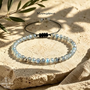 Può includere: Un braccialetto di perline con pietre di luna sfaccettate e traslucide dal sottile riflesso blu, arricchito da piccoli distanziatori in metallo argentato. Il bracciale presenta un cordino nero con chiusura a nodo scorrevole e il testo Moonstone.