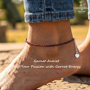 May include: A garnet anklet featuring small, deep red, round gemstone beads strung on a delicate cord. The anklet includes a silver-toned metal chain extension with a small heart-shaped charm at the end. Text reads: Garnet Anklet, Fuel Your Passion with Garnet Energy.