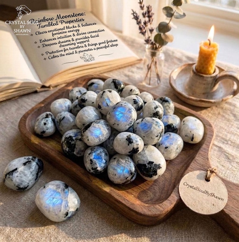 Pu&ograve; includere: Una collezione di pietre burattate in pietra di luna arcobaleno lucide con inclusioni bianche, grigie e nere, caratterizzate da riflessi blu iridescenti. Le pietre sono esposte su un vassoio di legno con un'etichetta rotonda CrystalSoulByShawn.