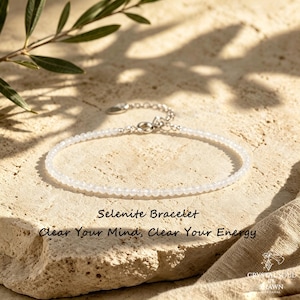 Può includere: Un braccialetto delicato con piccole perline di selenite bianca traslucida infilate su un filo sottile con una chiusura in metallo color argento e una catenella di estensione. Il testo "Selenite Bracelet" e "Clear Your Mind, Clear Your Energy" è stampato.