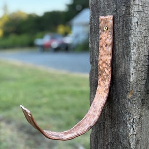 May include: A handcrafted copper hook mounted on a weathered wooden post. The hook has a textured surface and a curved design, ideal for hanging items. The background shows a blurred view of a road and greenery.