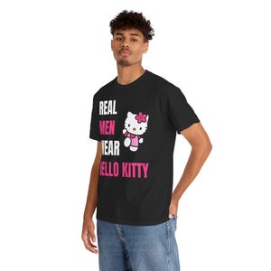 Może przedstawiać: Czarny t-shirt z napisem "REAL MEN WEAR HELLO KITTY" w kolorze białym i r&oacute;żowym. Koszulka przedstawia grafikę Hello Kitty w r&oacute;żowej sukience i kokardce. Model ma na sobie niebieskie dżinsy.