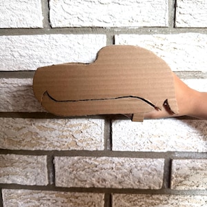 Puede incluir: Un recorte de cartón marrón con forma de perfil lateral de un coche, que presenta una línea negra dibujada a mano que define el capó, el parabrisas y la línea del techo, con una pequeña pestaña rectangular que sobresale del borde.