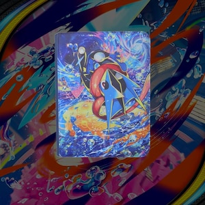 May include: A rectangular protective case featuring a vibrant, colorful illustration of a blue and black frog-like creature with a long pink tongue. The artwork includes swirling cosmic patterns, bubbles, and bright orange, blue, and purple hues.