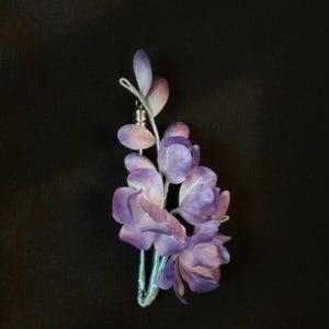 May include: A floral brooch featuring layered purple and light violet petals attached to a light blue stem. The piece includes a silver-toned metal pin mechanism and displays the text Minglienne x Obliquetable at the bottom of the frame.