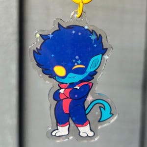 May include: A clear acrylic keychain featuring a cartoon character with blue skin, dark blue hair, and a yellow eye patch. The character wears a dark blue and red suit with white boots and has a blue tail. It includes a yellow star-shaped clasp.