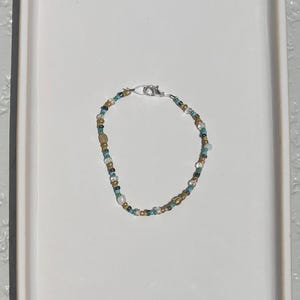May include: A beaded bracelet featuring a variety of small, multi-colored glass and metallic beads in shades of turquoise, brown, gold, and clear. The bracelet is secured with a silver-toned metal lobster claw clasp and jump ring closure.