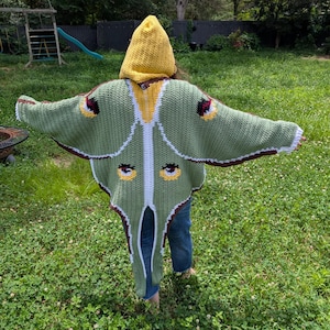 May include: A crochet hooded cloak designed to resemble a luna moth, featuring a yellow hood, sage green wings with white trim, and dark brown edges. The garment displays four circular eye-like patterns in yellow, black, and white on the wings.