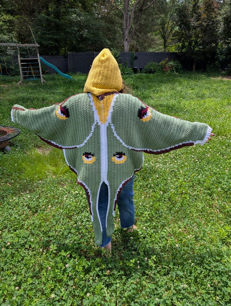 May include: A crochet hooded cloak designed to resemble a luna moth, featuring a yellow hood, sage green wings with white trim, and dark brown edges. The garment displays four circular eye-like patterns in yellow, black, and white on the wings.