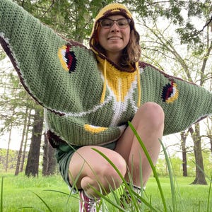 May include: A handmade crochet hooded sweater designed to resemble a moth, featuring wide, wing-like sleeves in sage green with white and yellow accents. The garment includes dark brown trim, a yellow hood, and eye-like patterns on the sleeves.