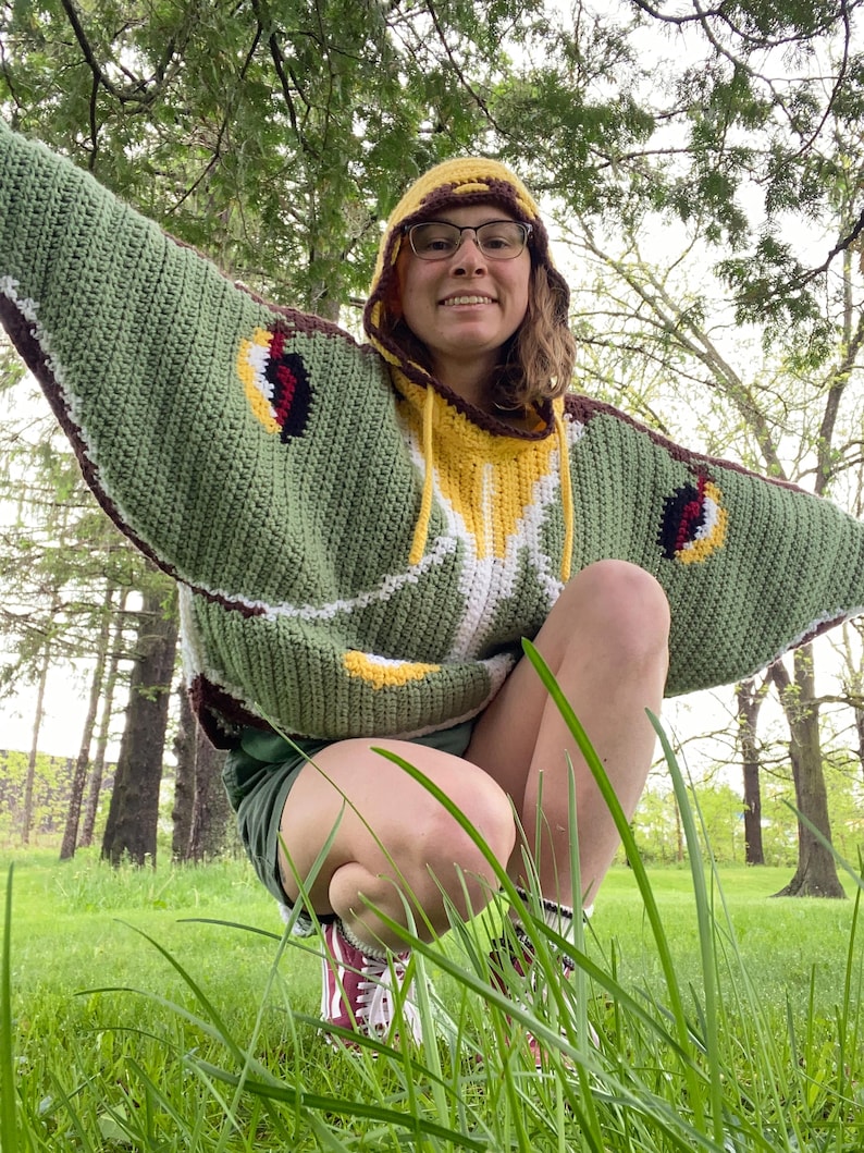 May include: A handmade crochet hooded sweater designed to resemble a moth, featuring wide, wing-like sleeves in sage green with white and yellow accents. The garment includes dark brown trim, a yellow hood, and eye-like patterns on the sleeves.