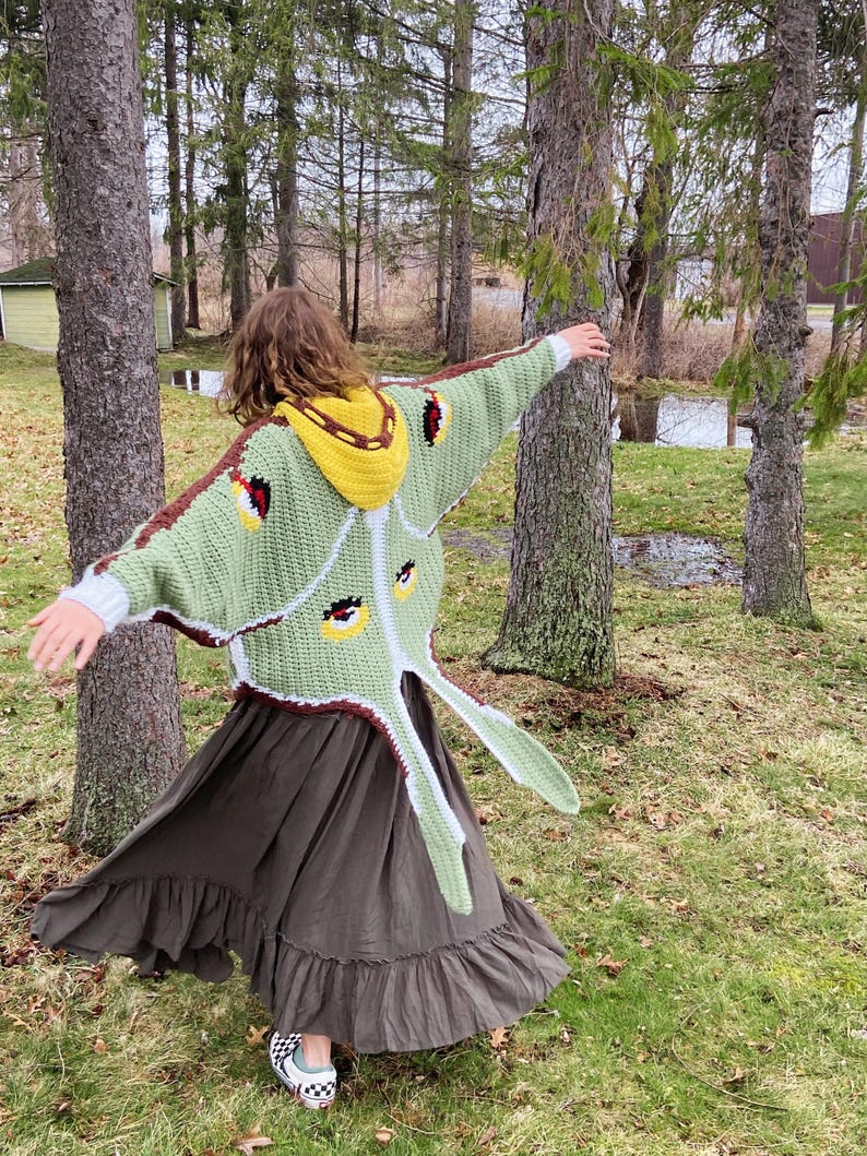 May include: A crochet cardigan designed to resemble a moth with a light green body, dark brown trim, and a yellow hood. The back features large wing-like panels with eye-spot patterns in yellow, black, and red, paired with a dark brown skirt.