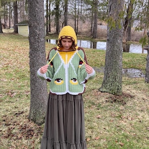 May include: A crochet hooded sweater featuring a light green body with white trim, a yellow hood, and brown accents. The sweater displays two stylized eye motifs on the front and includes long yellow drawstrings hanging from the hood neckline.