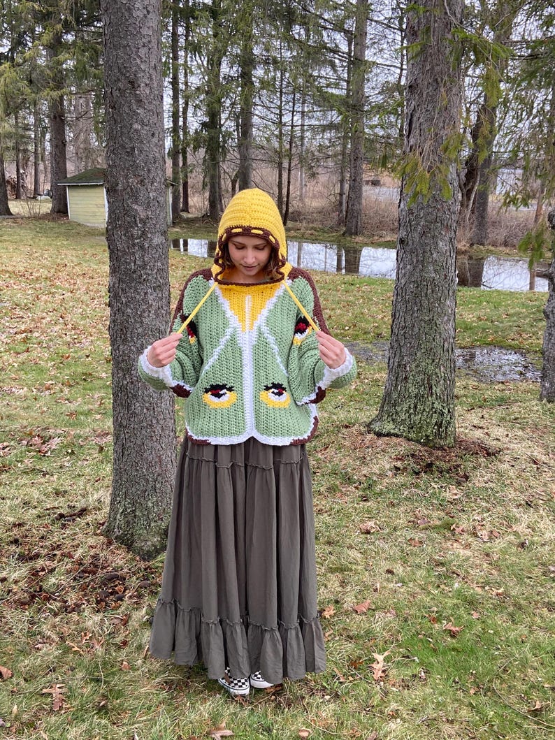 May include: A crochet hooded sweater featuring a light green body with white trim, a yellow hood, and brown accents. The sweater displays two stylized eye motifs on the front and includes long yellow drawstrings hanging from the hood neckline.