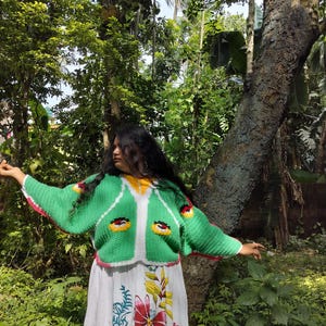 May include: A bright green crochet cardigan featuring a white front border, yellow neckline, and stylized eye motifs on the chest, worn over a white skirt with a colorful floral print featuring red, yellow, and blue botanical patterns.