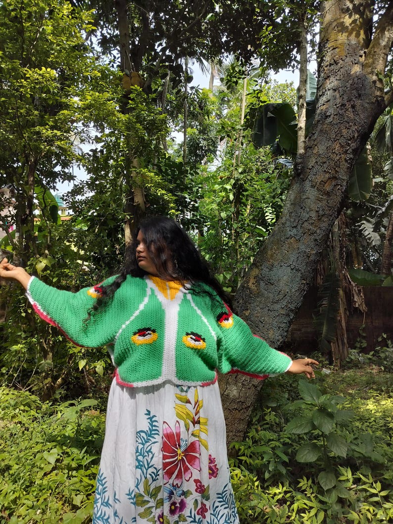 May include: A bright green crochet cardigan featuring a white front border, yellow neckline, and stylized eye motifs on the chest, worn over a white skirt with a colorful floral print featuring red, yellow, and blue botanical patterns.