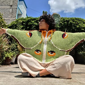 May include: A light green crochet zip-up hoodie featuring a moth wing pattern with dark brown, orange, and black circular eye-spot motifs on the sleeves and body, a white front zipper, and a yellow drawstring hood with a dark brown inner lining.