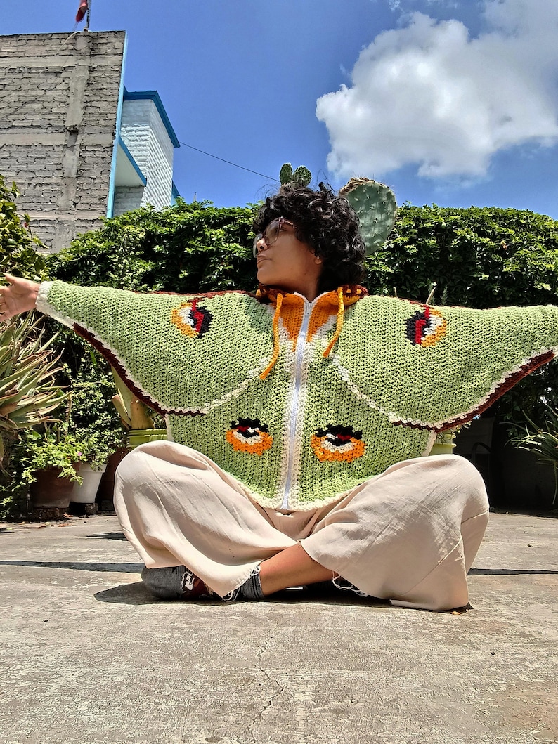 May include: A light green crochet zip-up hoodie featuring a moth wing pattern with dark brown, orange, and black circular eye-spot motifs on the sleeves and body, a white front zipper, and a yellow drawstring hood with a dark brown inner lining.