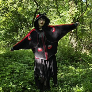 May include: A black crocheted hooded cardigan featuring wide bat-wing sleeves, a front zipper closure, and red accents along the shoulders and hem. The hood includes small black protrusions, and the text What in Yarnation is visible.