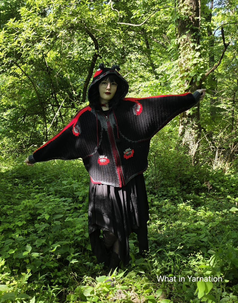 May include: A black crocheted hooded cardigan featuring wide bat-wing sleeves, a front zipper closure, and red accents along the shoulders and hem. The hood includes small black protrusions, and the text What in Yarnation is visible.